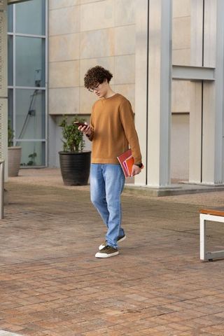 Male student walking campus courtyard checking phone carrying red folder wearing watch
