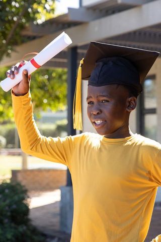 African american boy celebrating graduation holding diploma, wearing graduation cap in sunlight