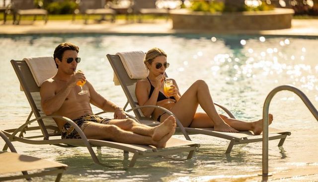 Sunlit resort couple relaxing on submerged loungers sipping orange cocktails
