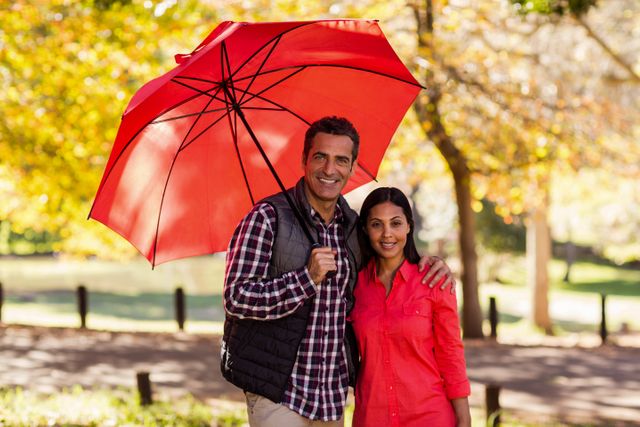 Couple standing under a red umbrella in a park during autumn. They are smiling and enjoying a sunny day surrounded by trees with fall foliage. Ideal for use in advertisements, relationship blogs, lifestyle magazines, and promotional materials for outdoor activities or romantic getaways.