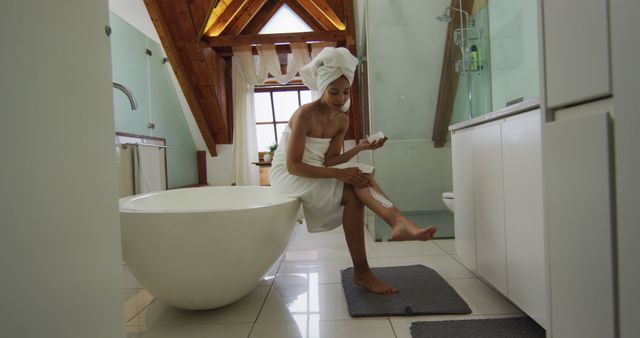 This stock photo features a woman applying lotion to her leg in a modern bathroom. She is wearing a towel wrap on both her body and head and is seated beside a freestanding bathtub. The bathroom has chic decor, and the ambiance suggests relaxation and self-care. Ideal for use in beauty and skincare promotions, home and lifestyle blogs, or wellness and self-care advertisements.