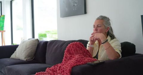 Mature woman sitting on sofa gazing thoughtfully under red throw in bright living room