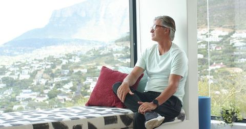 Mature man gazing through large window at mountain and coastal town in modern nook