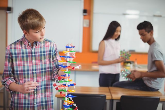 Schoolboy examining a colorful molecule model in a science class laboratory. Two other students are in the background, also working with molecular structures. Ideal for educational content, science and technology promotions, school brochures, and STEM learning resources.