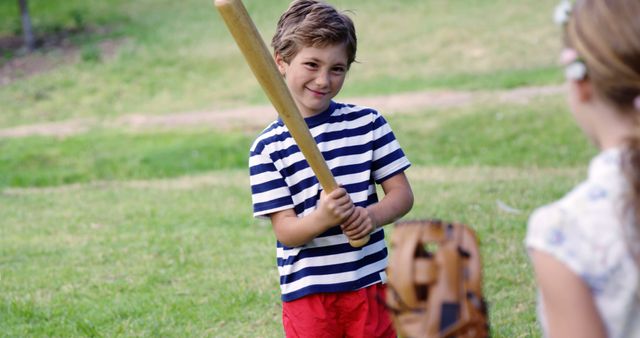 A young Caucasian boy holds a baseball bat with a smile, ready to play, as a girl with a glove prepares to catch, with copy space. They are enjoying a casual game of baseball in a grassy outdoor area.