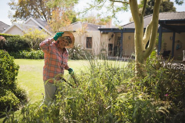 Senior woman enjoying gardening in her yard on a sunny day. She is wearing a hat and gloves, surrounded by lush greenery. Ideal for use in articles or advertisements related to active retirement, gardening tips, outdoor activities for seniors, or healthy lifestyle choices.
