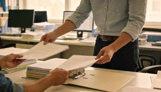 Office team exchanging paperwork over desk with open binder, stacked folders and monitors