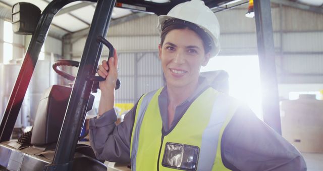 A young Caucasian woman in a hard hat and reflective vest stands confidently by a forklift, with copy space. Her attire and the industrial setting suggest she works in a warehouse or logistics environment.