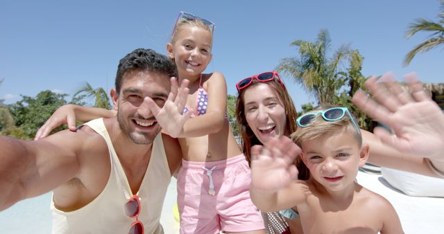 Family of four enjoying a beach vacation, waving at the camera with big smiles. Various palm trees and blue sky in background adding tropical vibe. Great for family travel advertisements, tourism brochures, vacation rentals, beachwear promotions.