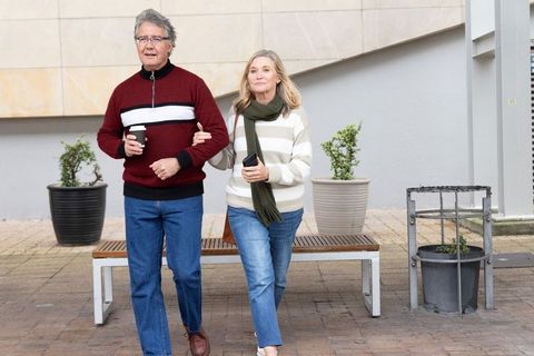 Senior couple walking arm-in-arm on urban plaza holding coffee and smartphone during casual outing