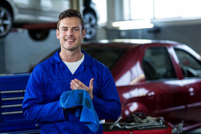 Mechanic in blue uniform smiling while wiping hands with cleaning cloth in an auto repair garage. Ideal for use in automotive service advertisements, repair shop promotions, and articles on car maintenance and professional services.