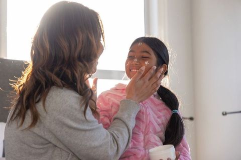 Mother applying moisturizer on daughter wearing pink robe, backlit tender morning bond