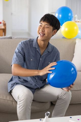 Smiling asian man sitting on sofa holding blue balloon celebrating casual home party