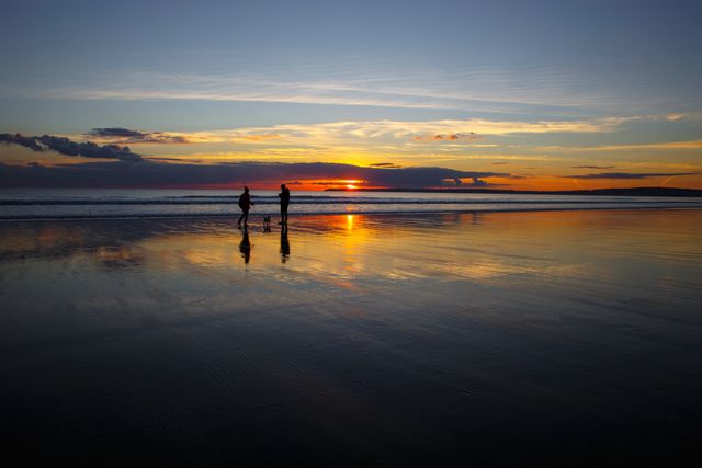 Family taking a walk on a beach during sunset. Their silhouettes are reflected on the wet sand, and the vibrant colors of the sunset sky create a serene atmosphere. This scene evokes feelings of peace, bonding, and relaxation, making it suitable for use in travel brochures, family-oriented advertisements, or any creative project related to nature and outdoor activities.
