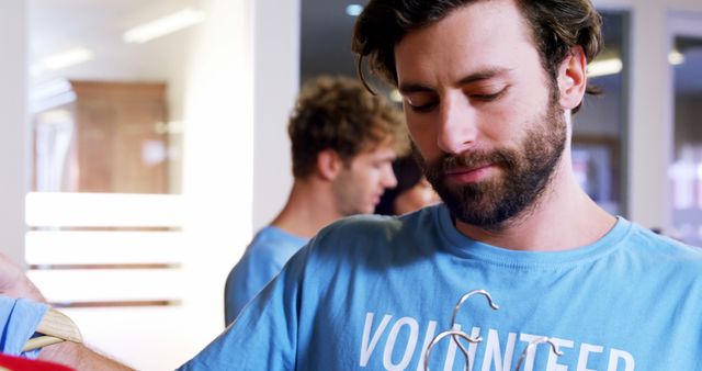 Young man with beard wearing blue volunteer shirt organizing clothes on hangers. Ideal for illustrating community service, charitable activities, volunteer recruitment, and social responsibility initiatives. Perfect for use in social media posts, websites, brochures, and advertisements related to volunteering and community charity events.