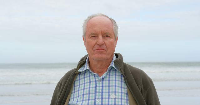 Elderly man standing by the ocean in casual clothing with a serious expression. Ideal for depicting senior lifestyle, outdoor activities, retirement themes, and introspective moments.