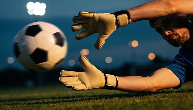 Goalkeeper diving and reaching to catch soccer ball at dusk wearing tan gloves and blue jersey
