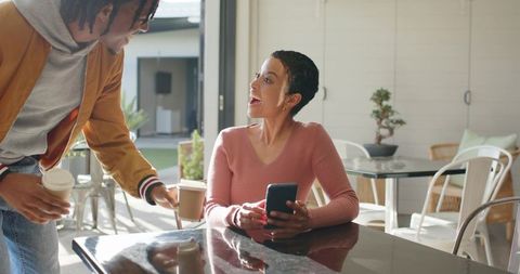 Friends sharing coffee on sunlit cafe patio casual meet woman holding smartphone laughing