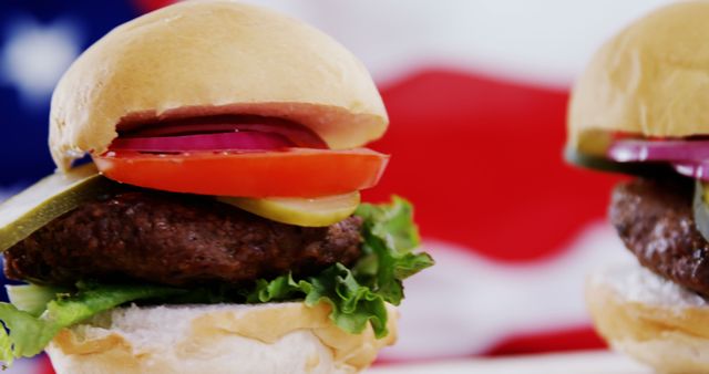 Close-up of mouthwatering American burgers adorned with layers of fresh lettuce, red tomato, onion, and pickles against a blurred background resembling the American flag. Ideal for use in advertisements for burger joints or fast food restaurants, food blogs, BBQ party promotions, social media food posts, and culinary websites.