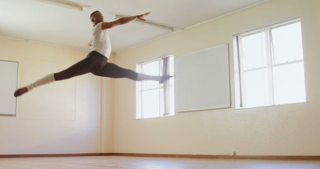 Male ballet dancer performing a high leap in a bright, spacious dance studio. Demonstrating athleticism, grace, and strength, the dancer exhibits skill and dedication to the art form. Ideal for content about professional dance training, athletic performance, male dancers, and ballet as a demanding art.