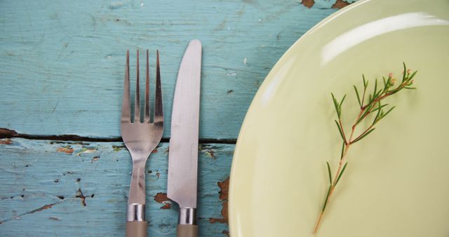 A fork and knife are placed beside a green plate with a sprig of rosemary on a rustic blue wooden table, with copy space. The setting suggests a preparation for a meal, emphasizing a simple and natural dining aesthetic.