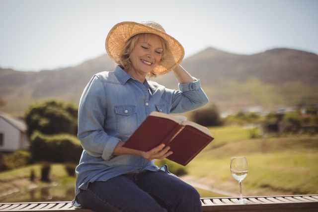 Senior woman sitting on park bench, enjoying sunny day. Wearing casual clothes and straw hat, holding book. Wine glass placed on bench. Ideal for themes like relaxation, retirement, outdoor leisure, peaceful living, reading habits, senior lifestyle.