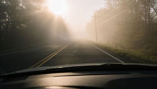 Driving into morning fog on rural two-lane road with sun glare and moody dash view