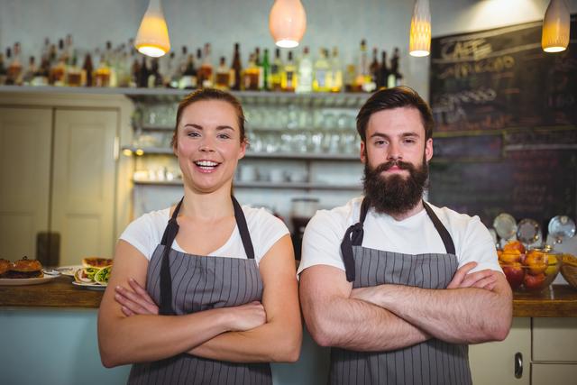 Waiter and waitress standing confidently at cafe counter, both wearing aprons and smiling. Ideal for use in articles or advertisements related to hospitality, customer service, teamwork, and restaurant business.