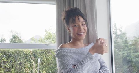 Cozy morning smiling woman in gray off-shoulder sweater holding ceramic mug by sunlit window