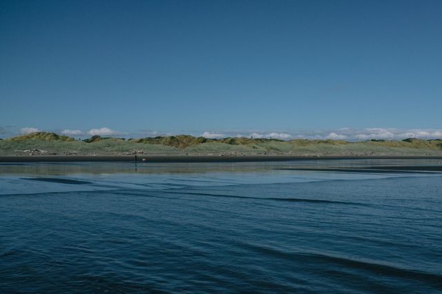 This image depicts a tranquil coastal landscape with a calm sea under a clear blue sky. Sand dunes stretch along the horizon, providing a serene and peaceful natural scene. This scene is perfect for backgrounds in travel and nature publications, promotional content for beach destinations, or home decor prints seeking to evoke tranquility and natural beauty.