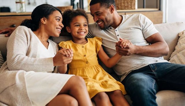 Smiling family holding hands on sofa, daughter in mustard dress sharing warm cozy moment
