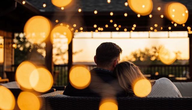 Couple snuggling on outdoor sofa under warm string lights at sunset romantic evening