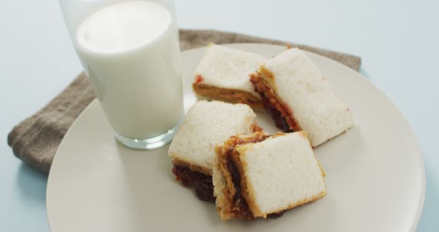Three peanut butter and jelly sandwiches served on a plate with a glass of milk on a light blue background. A beige napkin is placed beside the plate. Ideal for representing childhood snacks, lunch ideas, or a nutritious breakfast. Perfect for food blogs, recipe websites, and nutrition articles.