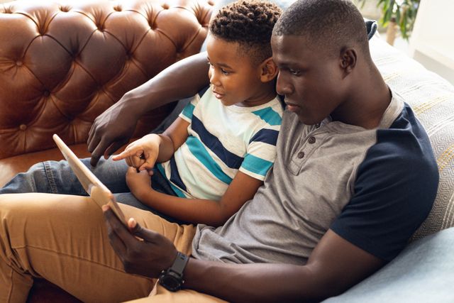 Father and son enjoying quality time together on couch, using a tablet. Perfect for themes of family bonding, parenting, technology use in families, and home life. Ideal for advertisements, blogs, and articles about family relationships, modern parenting, and digital learning.