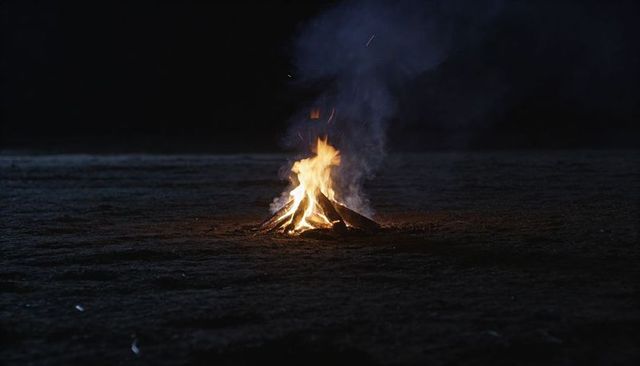 Solitary campfire burning on dark field at night with rising smoke and sparks