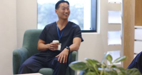 East asian male nurse relaxing in clinic breakroom holding coffee wearing scrubs smartwatch