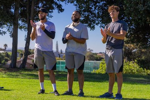 Diverse male teammates clapping and cheering on grassy park field during outdoor training