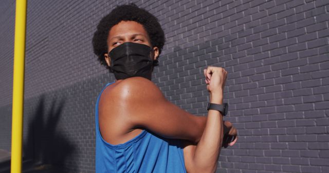 This image depicts a male athlete with afro hair stretching his arm outdoors while wearing a mask. He is dressed in athletic wear and is also wearing a wristwatch. Ideal for use in content related to fitness, outdoor exercise, health, and safety measures. Suitable for articles or campaigns focusing on physical training, sports, and pandemic precautions.