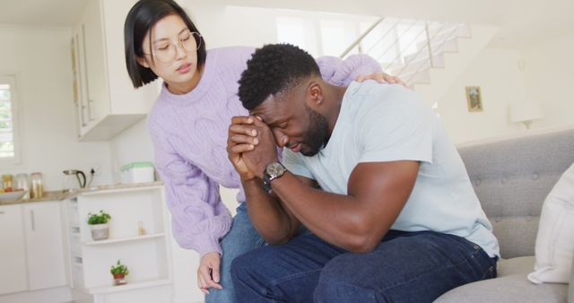 Female offering emotional support to distressed male in bright contemporary living room. Ideal for content on mental health, emotional support, compassion, and multicultural friendships.