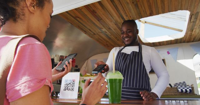 Happy female customer paying smiling african american male food truck owner by smartphone. small independent business, street food, service and catering.