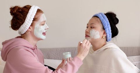 White and asian friends applying mint face mask together on sofa during cozy spa night