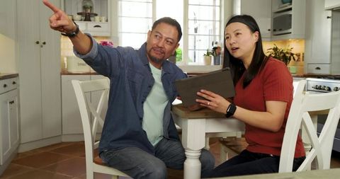 Asian Couple Planning Home Renovation at Kitchen Table Using Tablet and Pointing