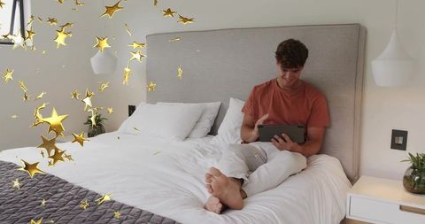 Young man relaxing on bed holding tablet while floating gold stars add magical tech vibe