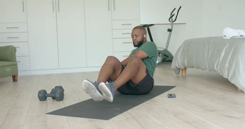 Man doing core workout on mat in minimal bedroom with dumbbell and exercise bike