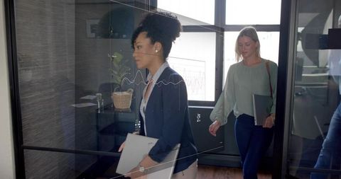Walking colleagues carrying laptops through glass-walled office with digital graph overlay