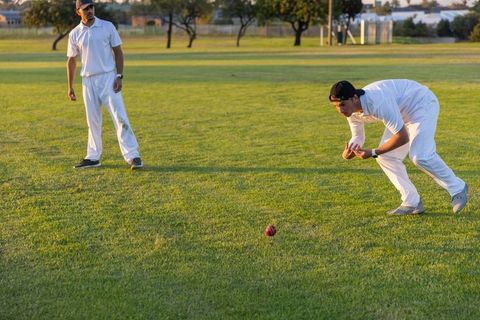 Diverse cricketers fielding on sunlit grass reaching for red ball during golden hour