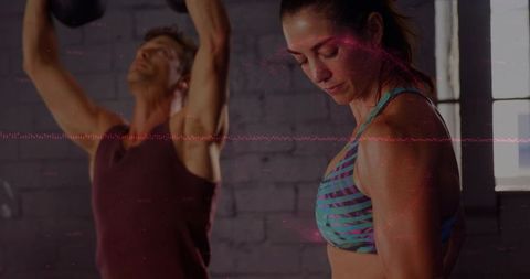 Woman checking form while partner lifts kettlebells in industrial loft gym daylight