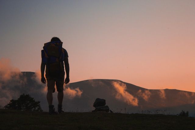 Silhouette of a solo hiker walking towards mountain range during sunset with backpack. Ideal for use in travel blogs, adventure promotions, nature magazines, outdoor brand advertisements, and inspirational motivational posts.