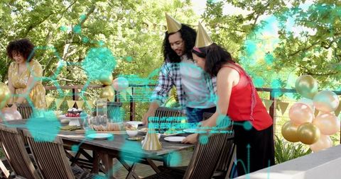 Friends arranging outdoor party table on wooden deck with balloons, party hats, festive decor