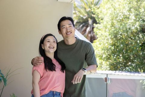 Asian couple smiling and looking up on balcony, embracing on sunny residential terrace